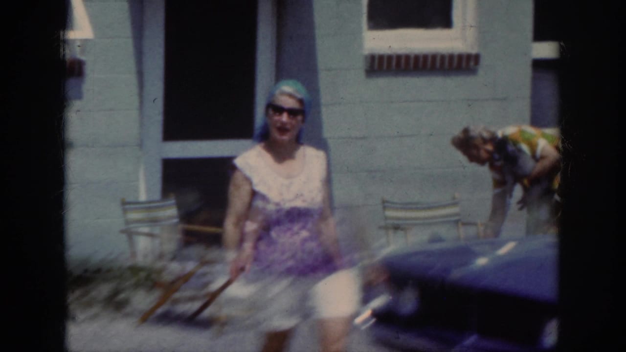 Woman approaches the camera beside a parked car while another adult bends near the building.