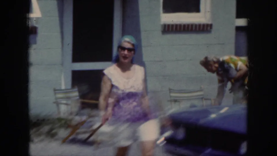 A woman in sunglasses and a blue head covering walks toward the camera beside a parked car in the stated 1962 Raleigh footage.