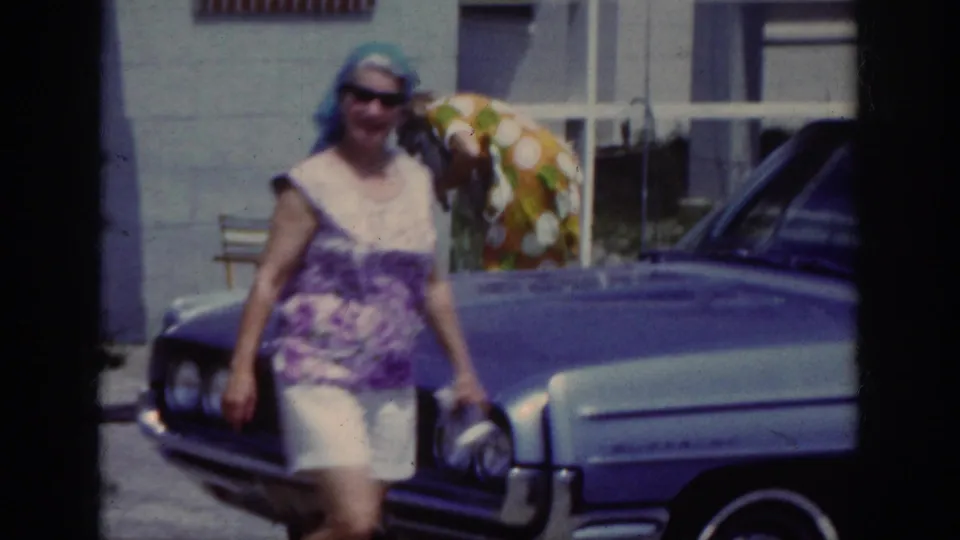 Close home-movie view of a woman in casual summer clothing walking past a blue car, with another adult visible behind her.
