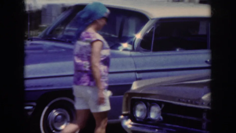 The woman is seen in profile walking between parked cars outside a light-colored building in the stated 1962 home movie.