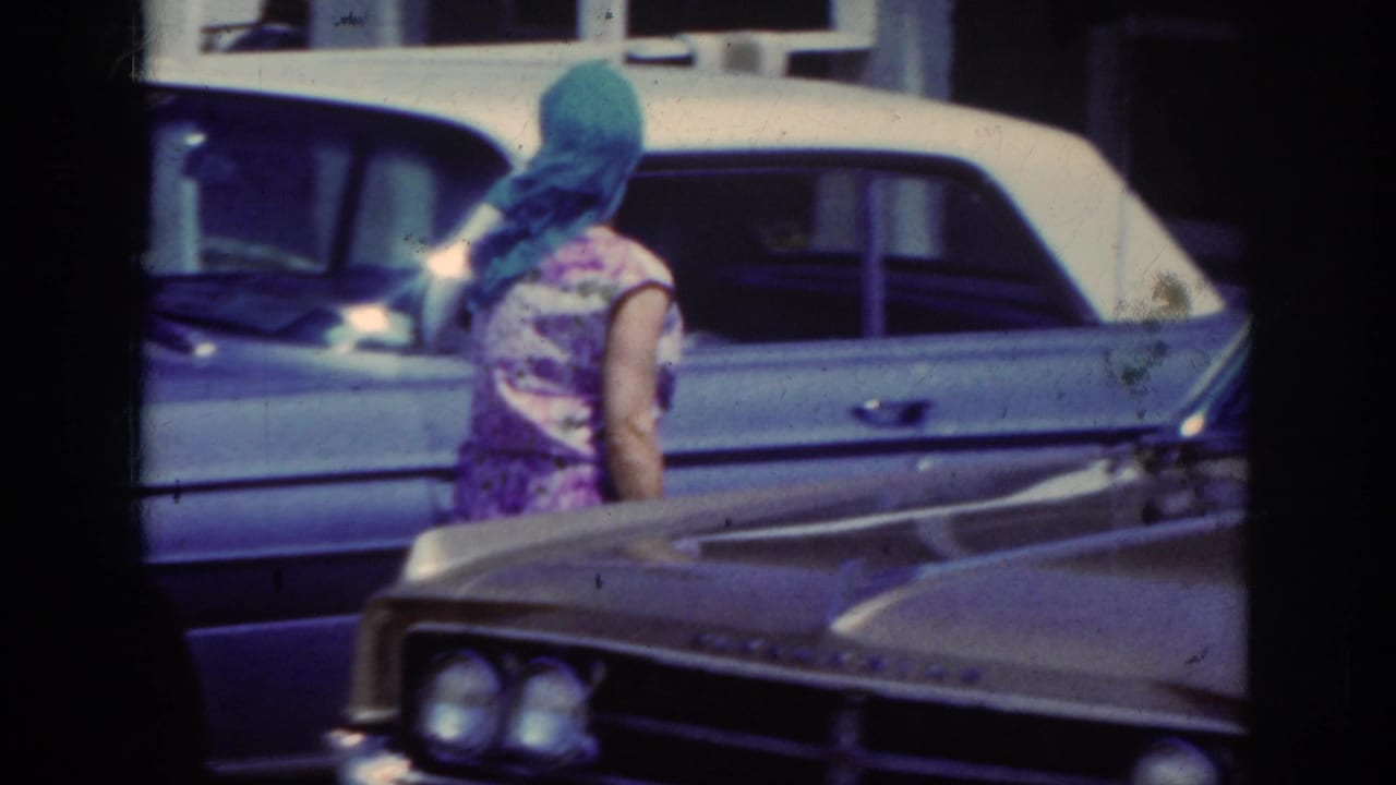 In 1962 home-movie footage, a woman in a floral dress and blue headscarf walks between parked older American cars.