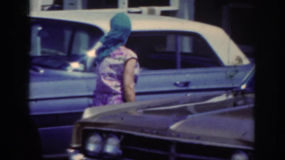 In 1962 home-movie footage, a woman in a floral dress and blue headscarf walks between parked older American cars.