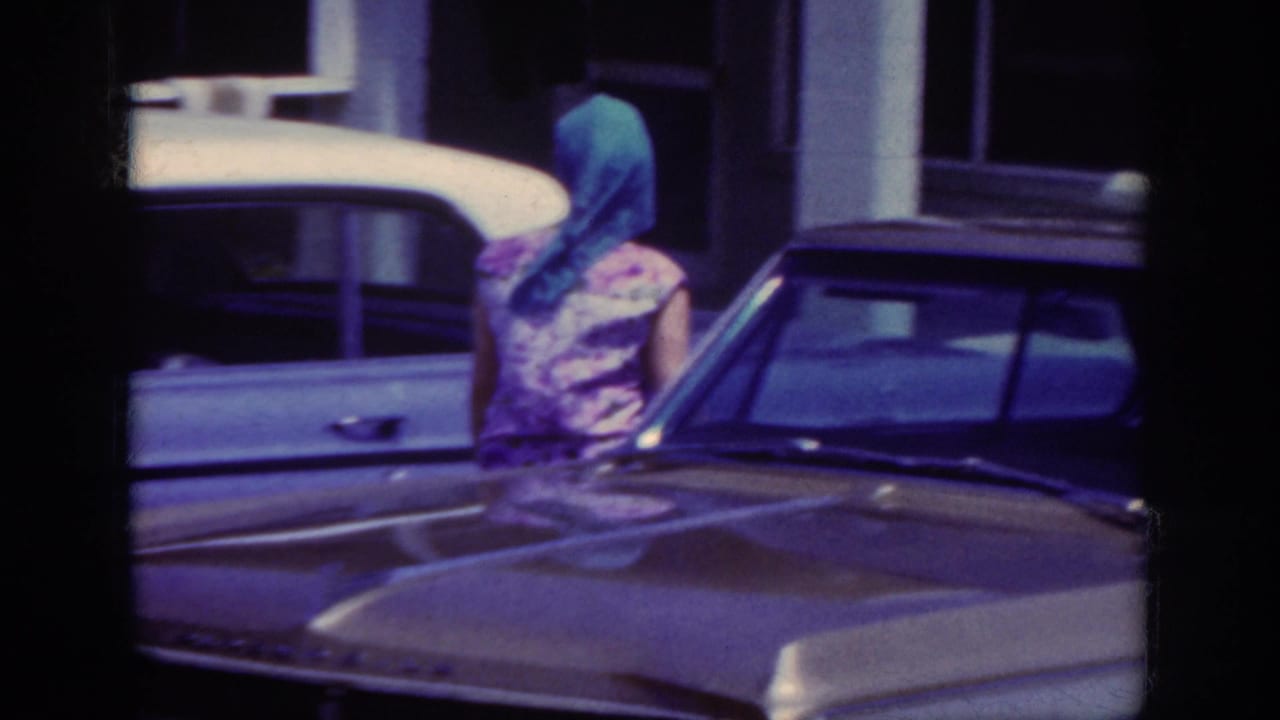 The woman in a blue headscarf stands or walks near a white sedan, with another parked car in the foreground.