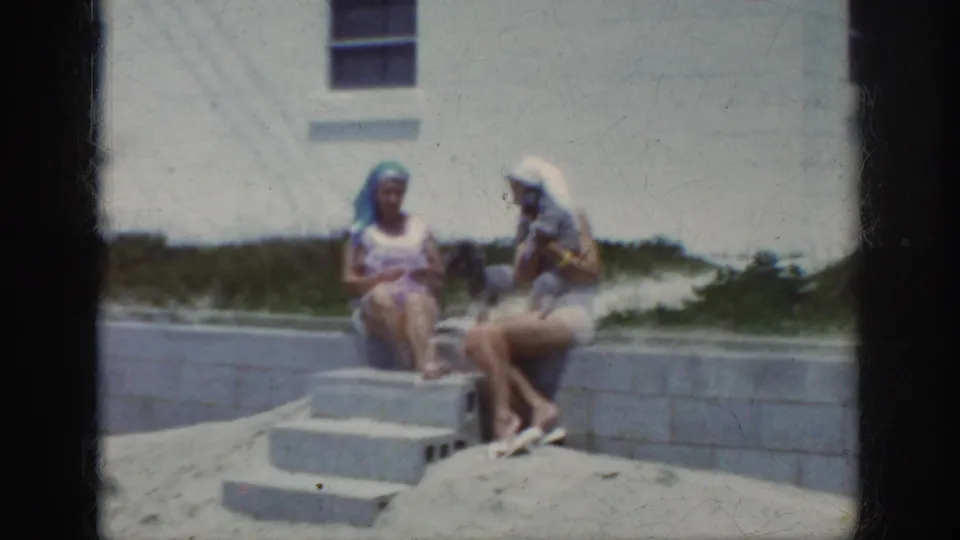 Two women in summer clothing sit on small concrete steps near sand and a pale wall in a beach-adjacent scene.