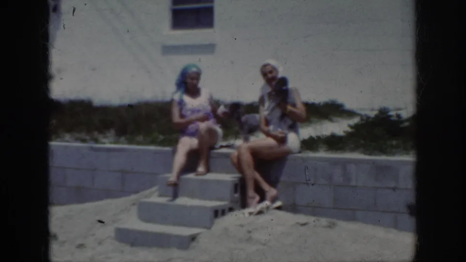 In 1962 home-movie footage, two women sit on concrete steps at a sandy beach area in Raleigh, North Carolina metadata context.