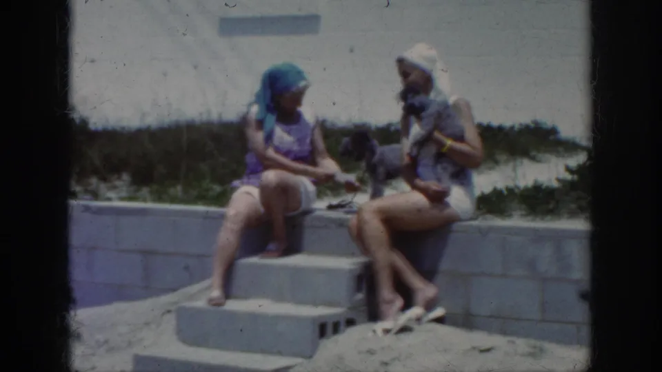 The women appear to talk on concrete beach steps while a small dog sits between them in the 8mm home-movie frame.