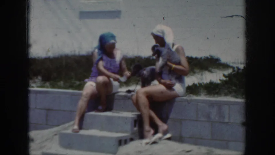 At the end of the clip, two women on beach steps look toward a small dog, with sand and a low wall behind them.