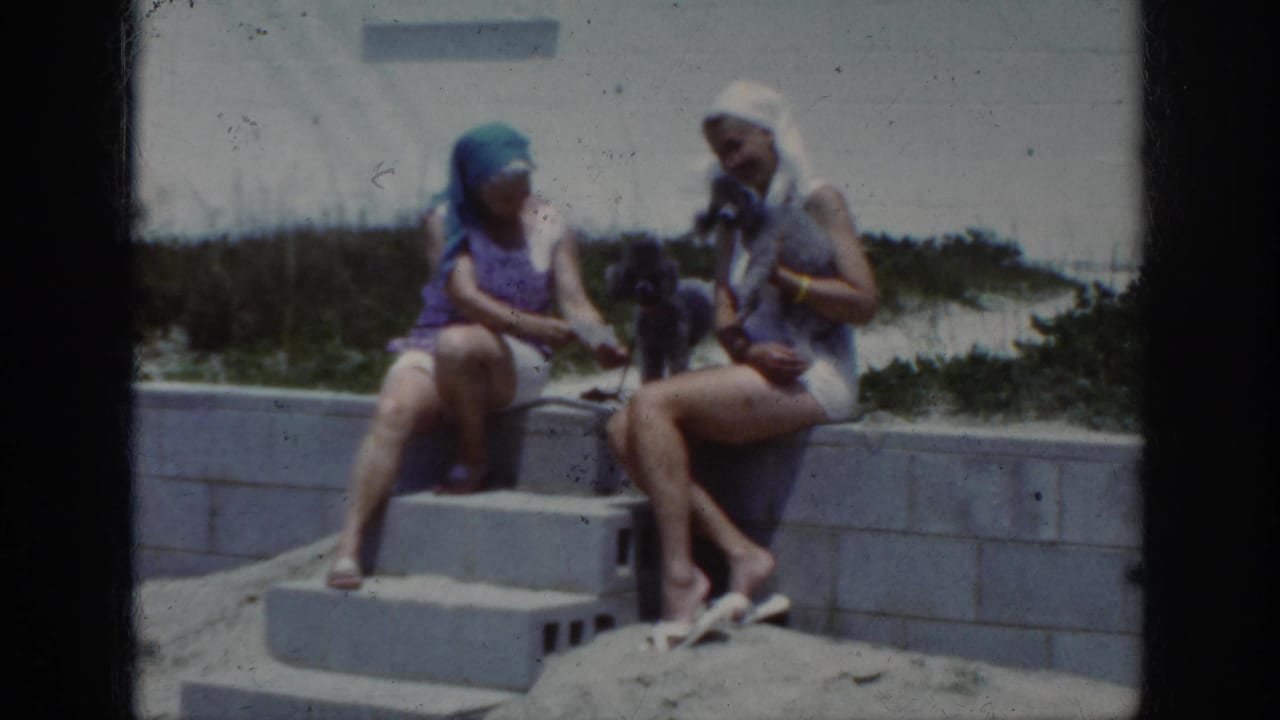 1962 home-movie frame of two women in swimsuits and head coverings sitting on beachside concrete steps with a small dark poodle.