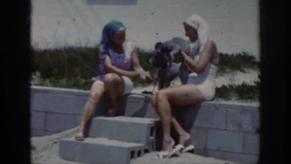 Beach home-movie frame showing one woman handling a small dark poodle as her seated companion reaches toward the dog.