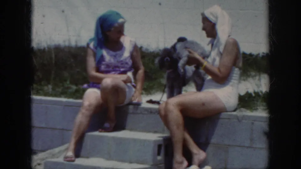 Two women in swimsuits and head coverings sit on beach steps in Raleigh metadata context, with a small dark poodle between them.