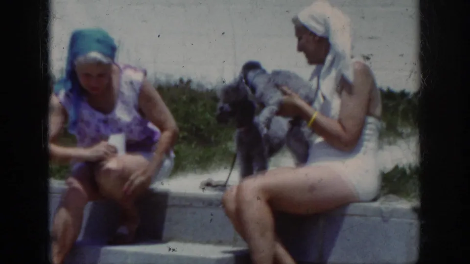1962 home-movie frame of two women in swimsuits and head coverings on concrete beach steps with a small dark dog.