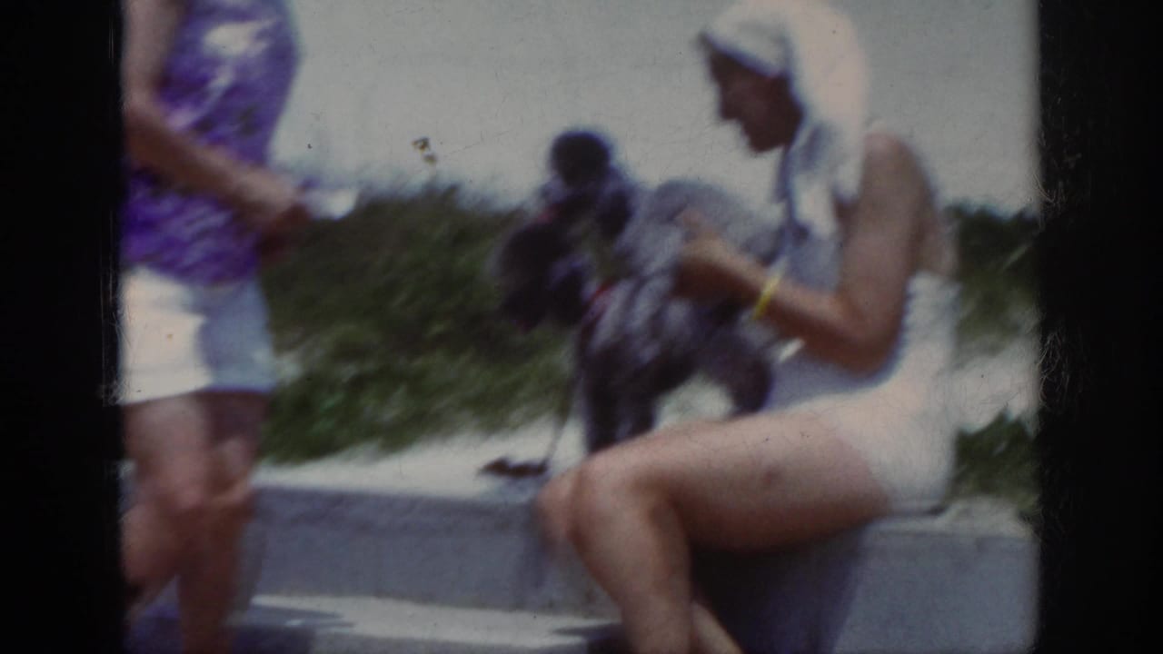 A woman in a white swimsuit sits on beach steps holding a small dog as another woman passes nearby.