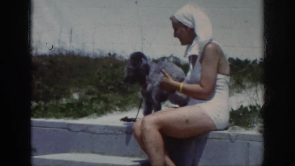1962 home-movie frame showing a woman in a white swimsuit and head covering holding a small dog on beach steps.