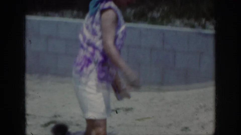 A woman in a purple patterned sleeveless top and white shorts walks across sand while a small poodle appears near the lower frame.
