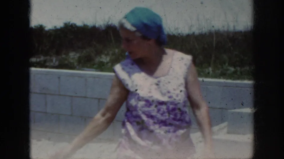 The woman, wearing a blue-green headscarf and purple patterned top, looks down and reaches near concrete steps in the 1962 home-movie scene.