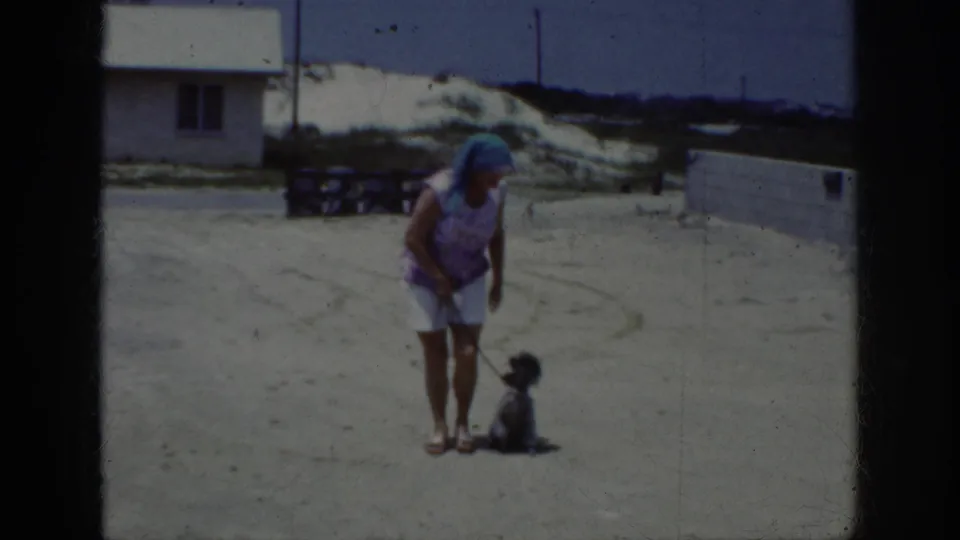 1962 Raleigh footage frame with a woman watching a small dark poodle sit on a leash near a white building and sandy ground.