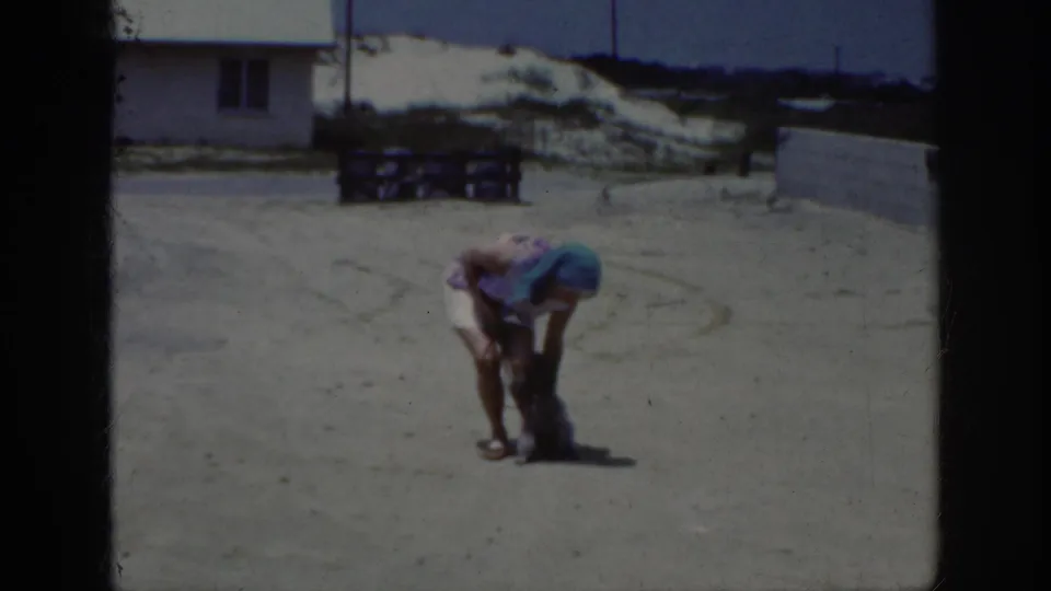 1962 Raleigh home-movie frame showing the woman bending down toward the small poodle as she adjusts or guides it during training.