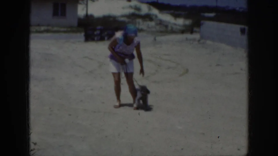 1962 Raleigh frame showing the woman standing near the small dark poodle after it sits, with sandy ground and low buildings behind them.