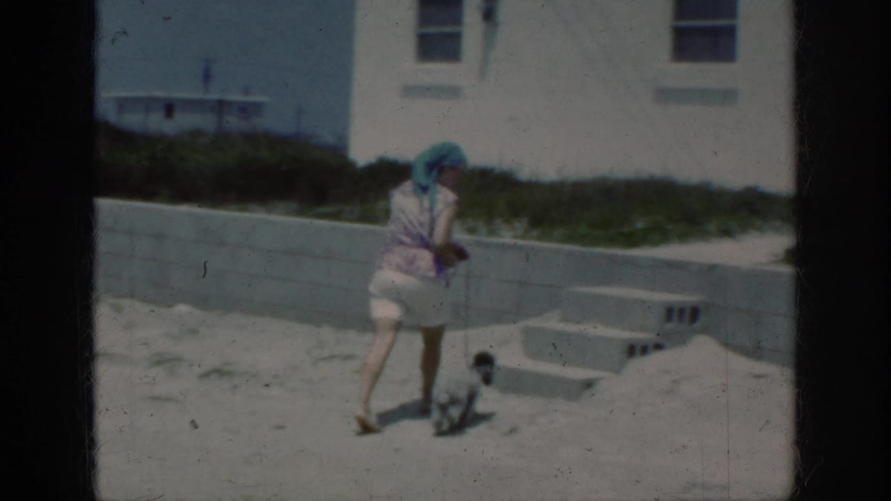 1962 8mm frame from Raleigh metadata showing a woman in headwear walking a small dog near sand, concrete steps, and a white building.