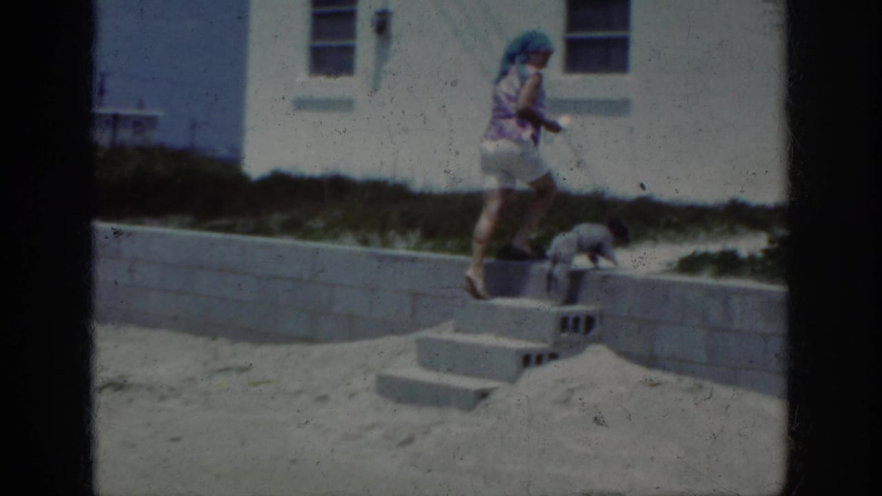 Woman steps along a low wall while a small dog walks near concrete steps on the sand.