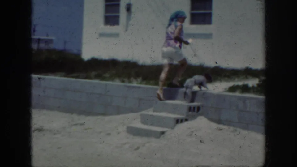 1962 Raleigh home-movie frame showing a woman in shorts and head covering walking a small dog beside steps and a white wall.