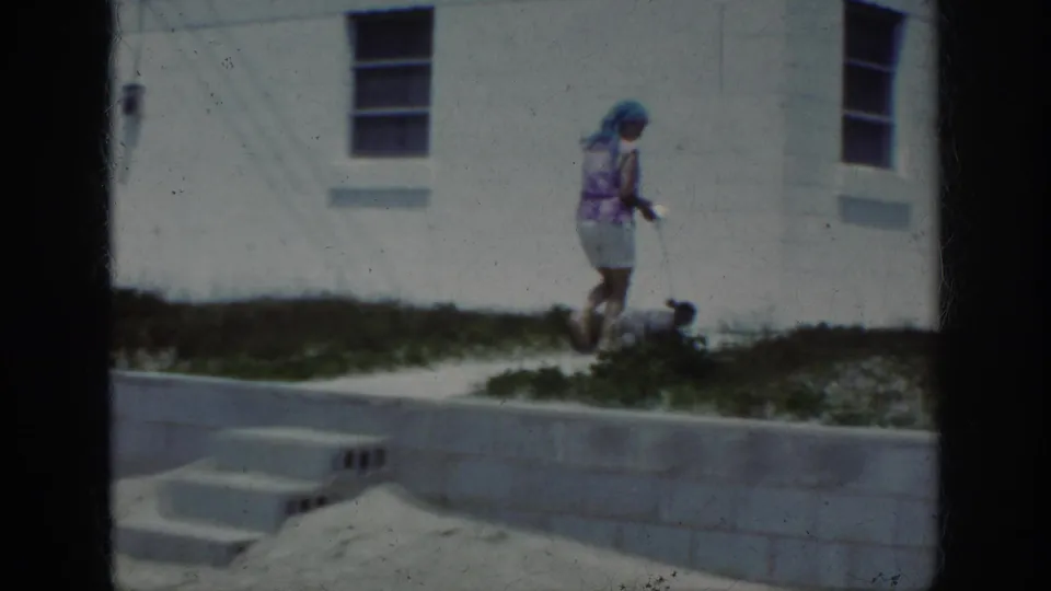 1962 8mm Raleigh frame with a woman walking a small dog near grass, sand, a retaining wall, and a white building facade.