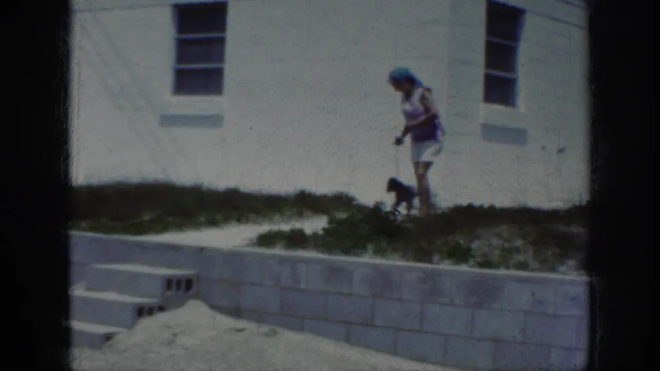 1962 Raleigh home-movie frame showing a woman attending to a small dog beside a low concrete wall, sand, grass, and a white building.