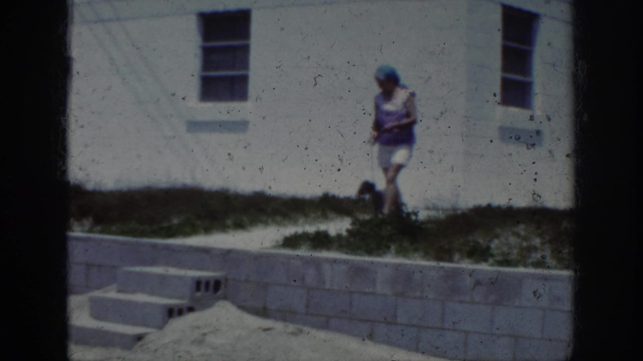 1962 home-movie frame of a woman walking a small leashed dog near a white wall, concrete path, steps, and sandy ground.