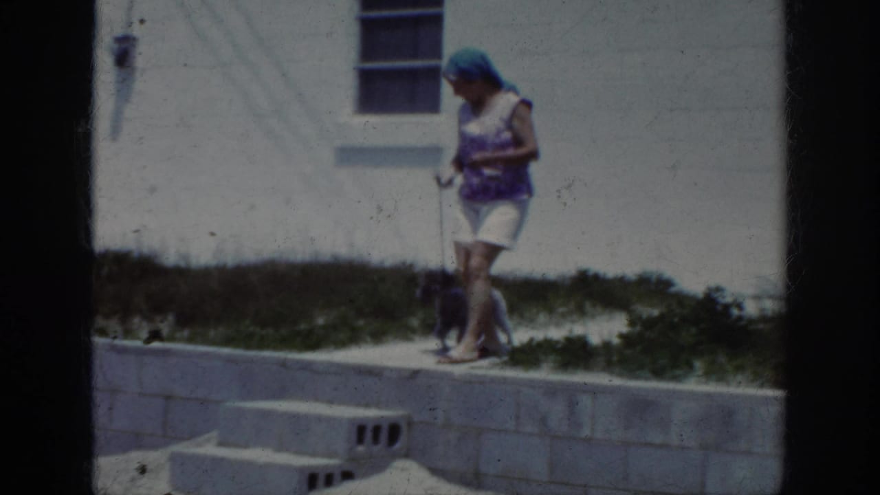 Woman in a purple top and white shorts walks barefoot with a small leashed dog near a white wall.