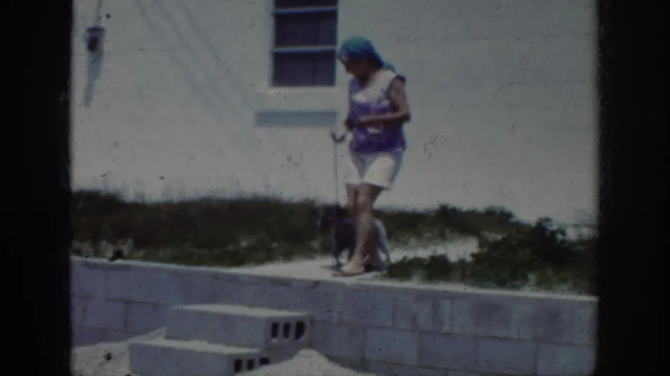 1962 home-movie frame showing the woman and small dog moving along a concrete edge beside a white building wall and window.