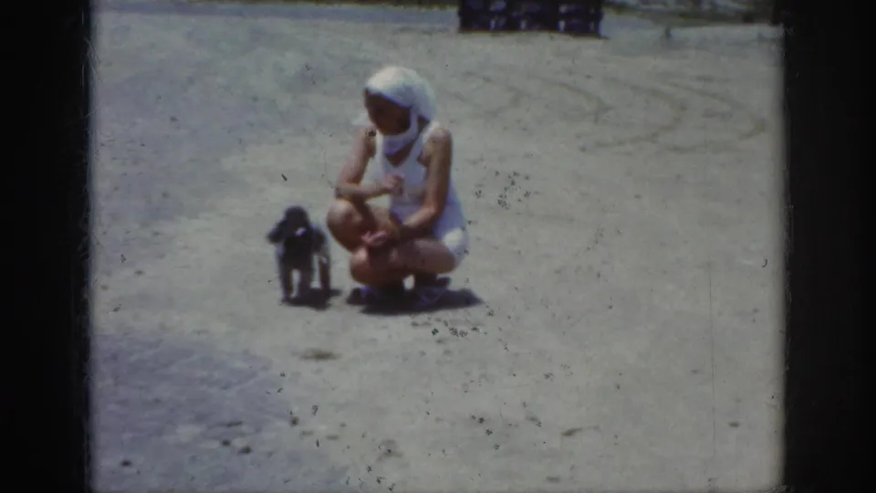 1962 Raleigh frame showing a woman in white clothing and a headscarf crouching beside a small dark poodle on sandy ground.