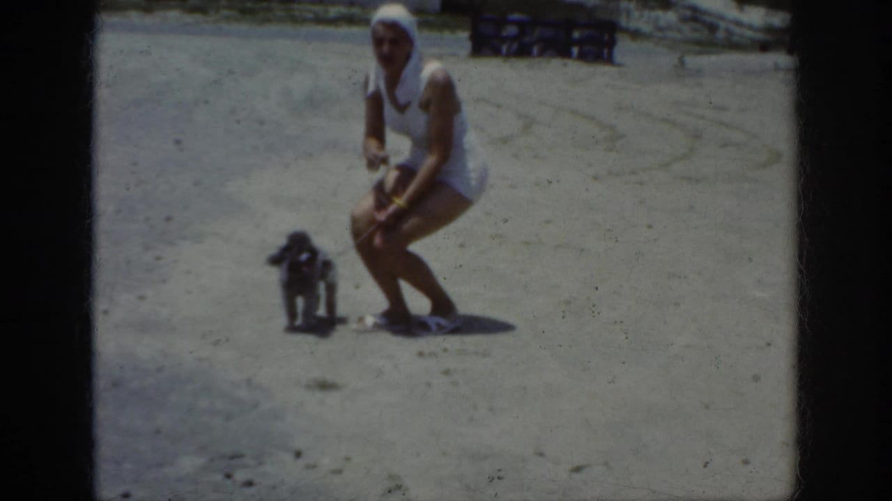 In 1962 home-movie footage, a woman in a white swimsuit and headscarf crouches beside a small dark dog on a sandy beach area.