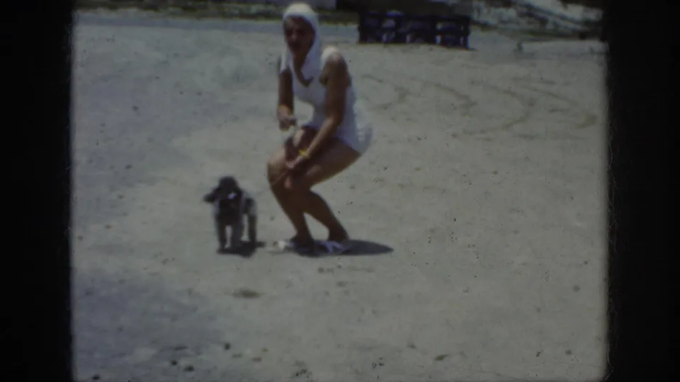 In 1962 home-movie footage, a woman in a white swimsuit and headscarf crouches beside a small dark dog on a sandy beach area.