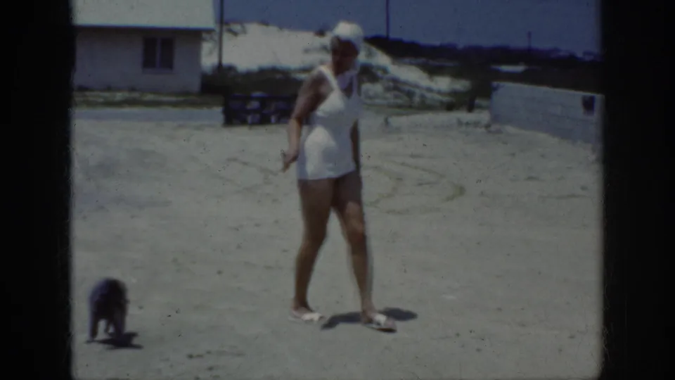 The woman pauses on the sandy beach area as the small dark dog moves near the lower edge of the home-movie frame.