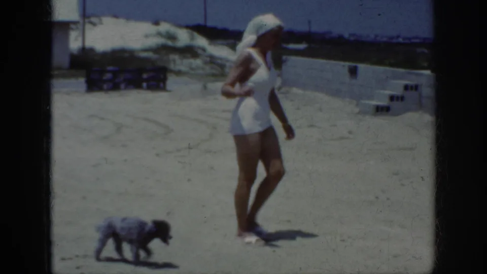 The woman walks across the sand in a white swimsuit and headscarf as the small dark dog crosses ahead, with low walls and dunes in the background.