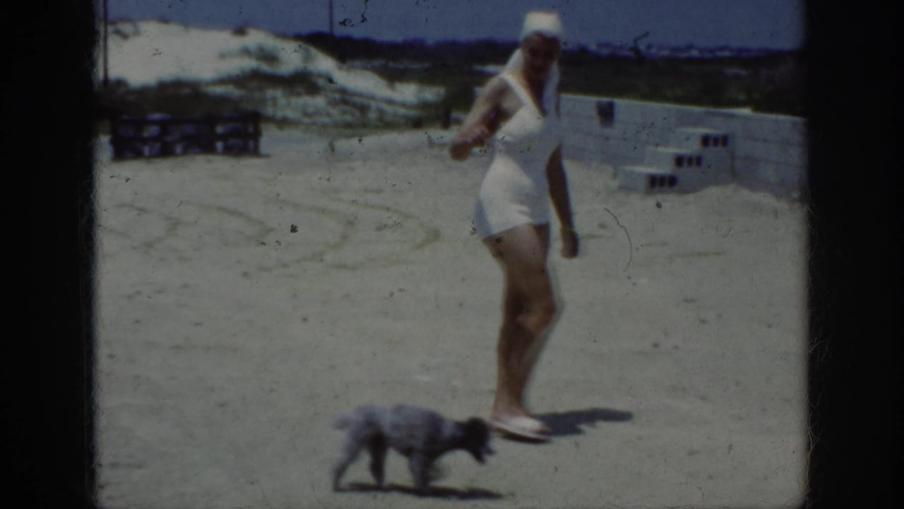 1962 Raleigh home-movie frame showing a woman in a white swimsuit and head covering walking two small dogs on a sandy terrace.