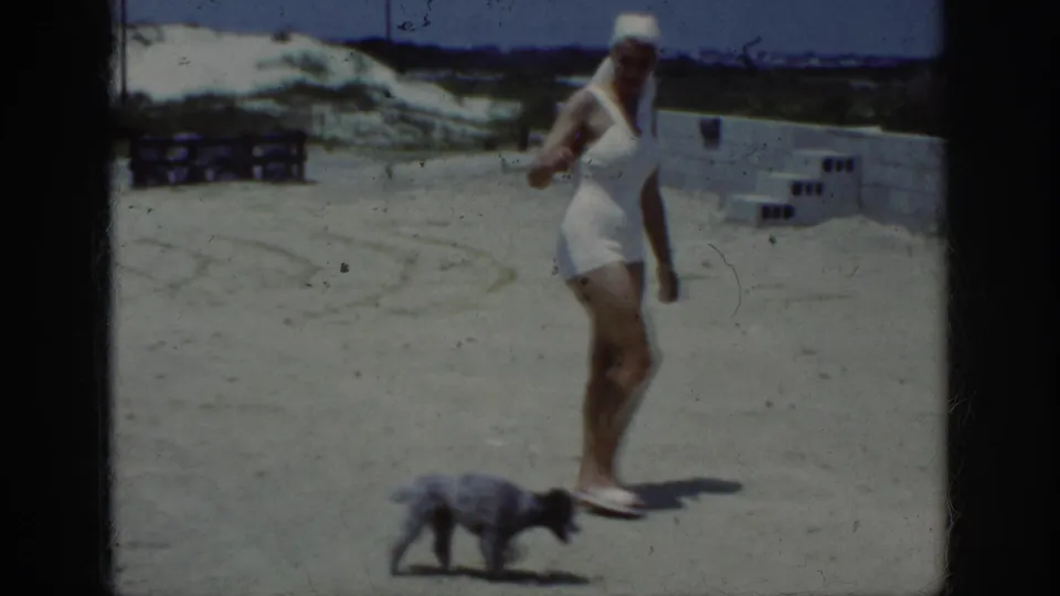 1962 Raleigh home-movie frame showing a woman in a white swimsuit and head covering walking two small dogs on a sandy terrace.
