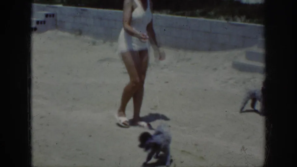 1962 Raleigh home-movie view of a woman walking on a sandy terrace as two small dogs move near the lower edge of the frame.