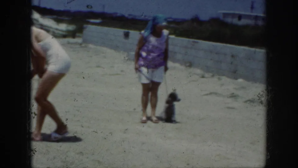 A woman in a purple patterned top and white shorts stands with a small poodle on sand in the 1962 Raleigh home-movie footage.