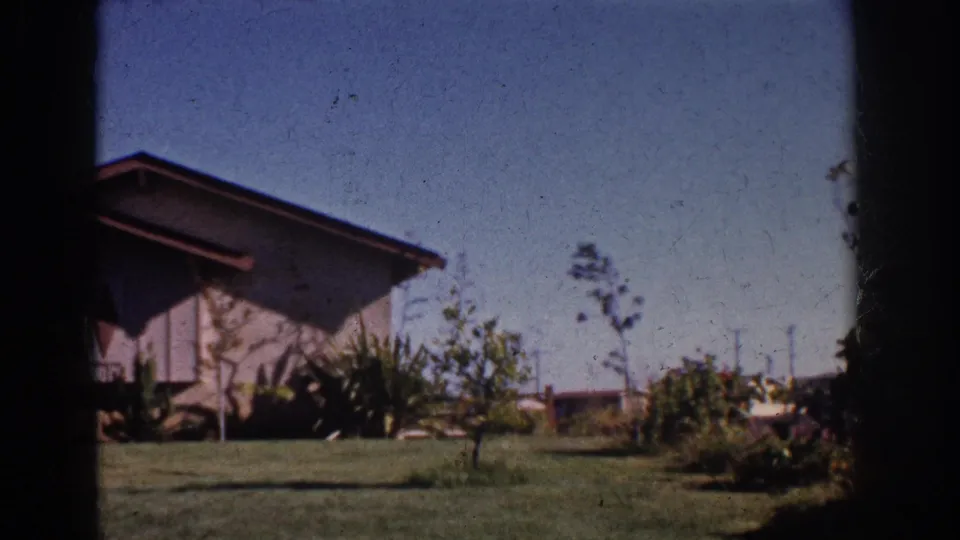 1962 home-movie frame showing the house corner, garden shrubs, trimmed grass, saplings, and blue sky in Yorba Linda.