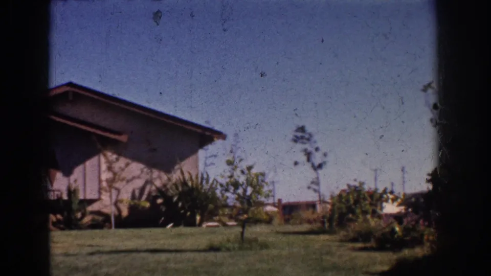 Yorba Linda front yard in 1962, with a sloped-roof house, low shrubs, sapling trees, lawn, and dark film border at the side.