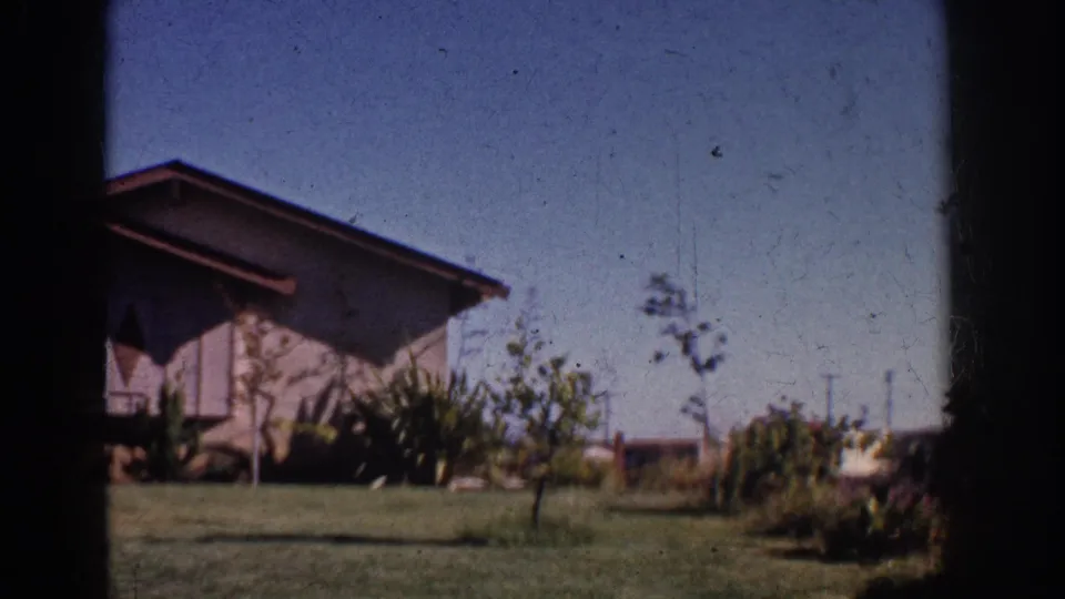 1962 Yorba Linda home-movie frame showing a mid-century house, lawn, small trees, and shrubs in bright daylight.