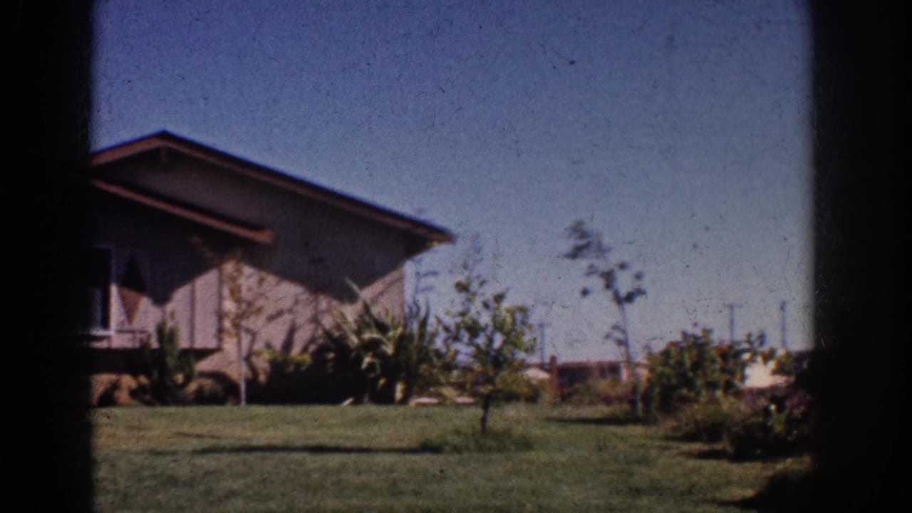 Suburban house partly framed by lawn, shrubs, small trees, and dark film edges.