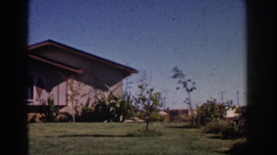 1962 Yorba Linda exterior view of a sloped-roof house with planted yard and wind-touched shrubs.