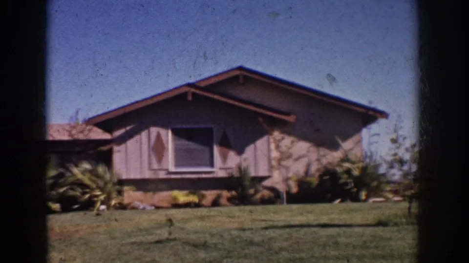 1962 Yorba Linda home-movie frame showing a mid-century ranch house, front lawn, shrubs, and palm-like landscaping under blue sky.
