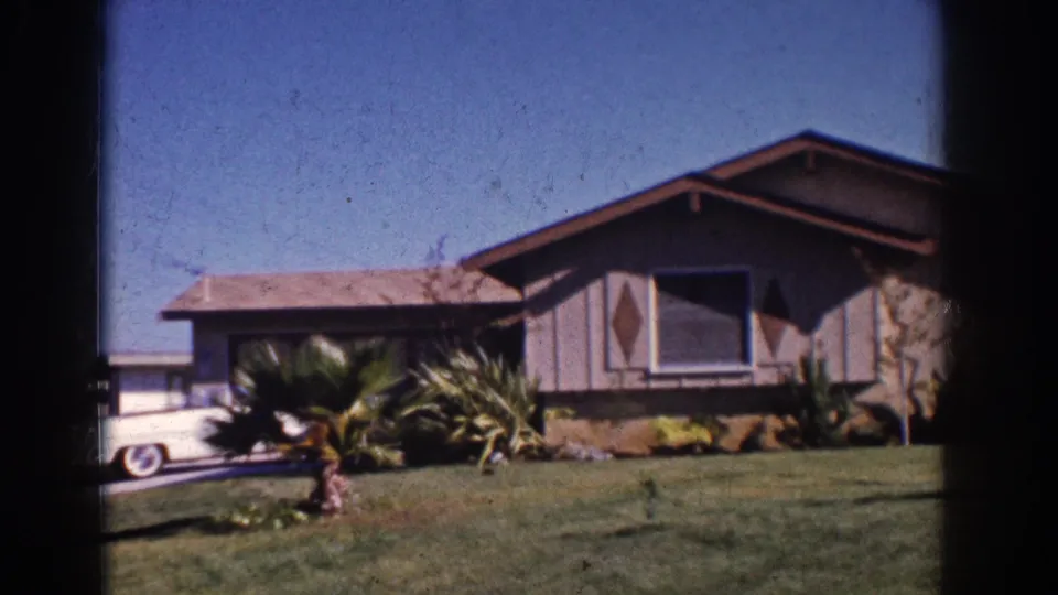 1962 Yorba Linda home-movie frame showing the ranch house, front lawn, landscaping, and part of a white 1960s sedan at left.