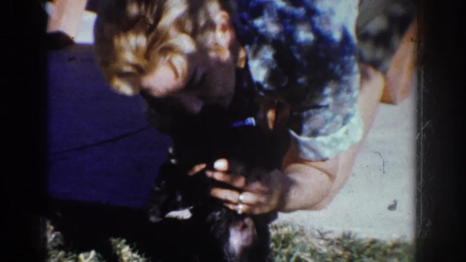 1962 Yorba Linda home-movie frame showing a person crouched over a small black puppy near a lawn and concrete sidewalk.