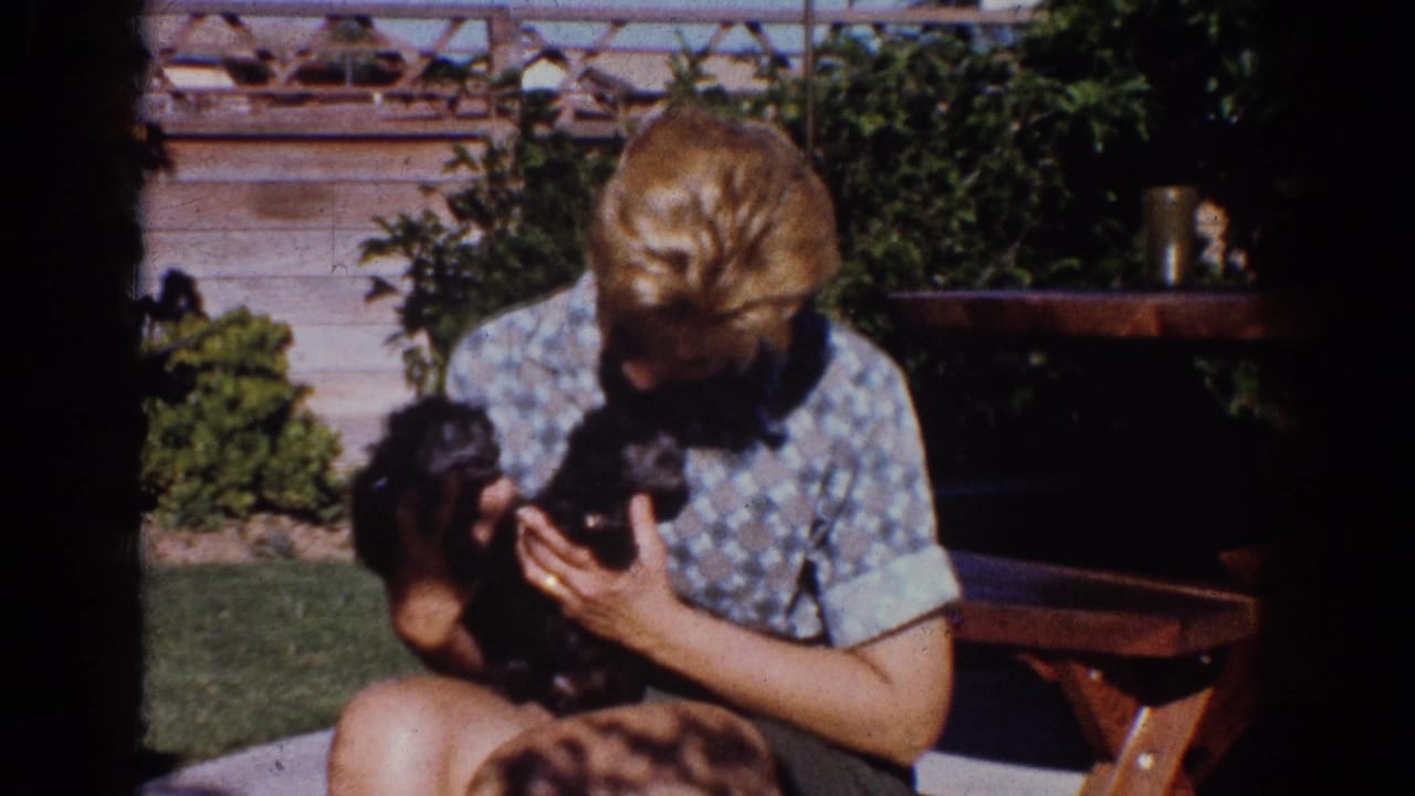 1962 Yorba Linda home-movie frame showing a young person in a patterned shirt cuddling a small black puppy in a backyard.