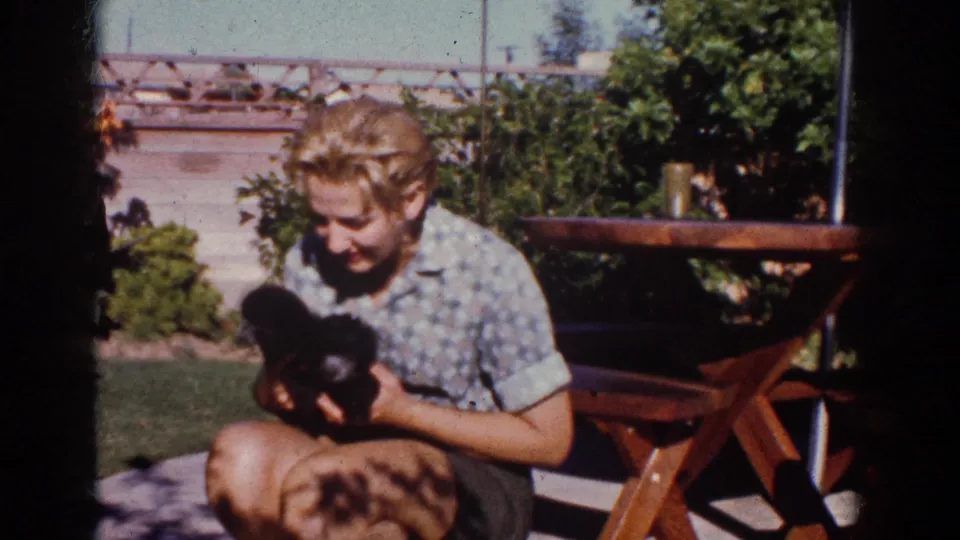 A smiling young person holds a small black puppy in a sunlit 1962 Yorba Linda backyard, with lawn and picnic table behind.