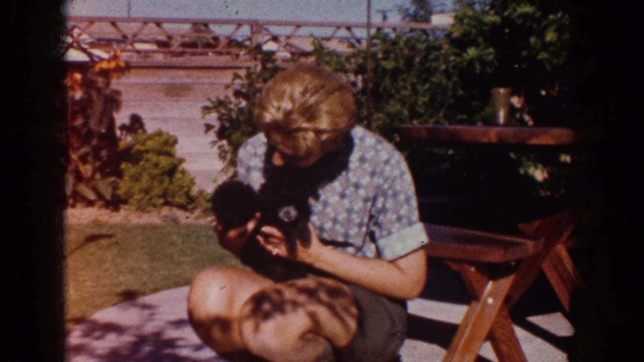 In Yorba Linda, California, 1962, a woman in a patterned blouse kneels on a lawn and holds a small black puppy beside a wooden picnic table.